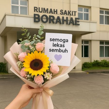 Tangan wanita memegang buket bunga dengan kartu bertuliskan “semoga lekas sembuh 💜” di depan Rumah Sakit